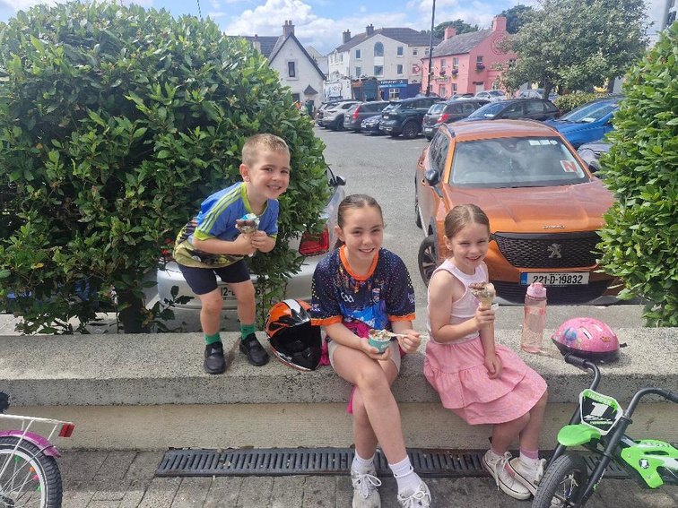 Andrea’s three children sitting together on a low wall on a sunny street, with their bikes and helmets nearby