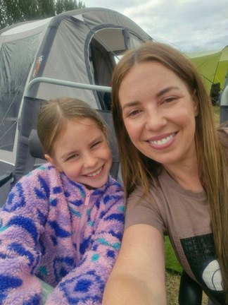 Andrea smiling beside her daughter in a colourful fleece outside a tent on a family camping trip