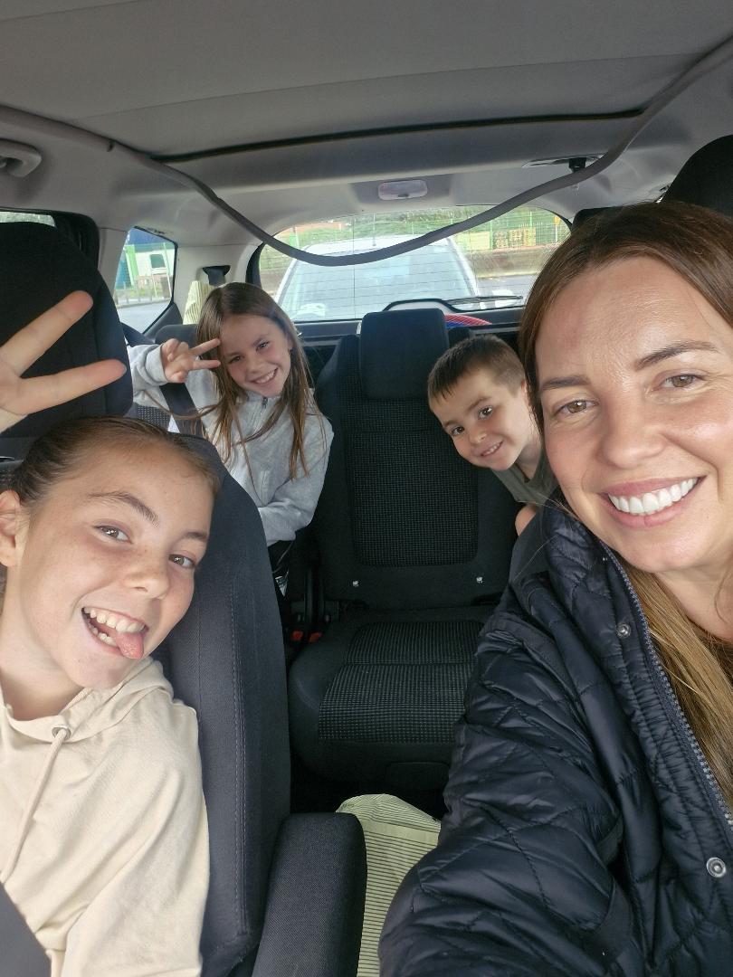 Andrea taking a cheerful car selfie with her three children, one pulling a funny face and another giving a peace sign